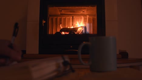 Fondo-Acogedor-De-Una-Joven-Mujer-Escribiendo-A-Mano-En-Un-Viejo-Cuaderno-Cerca-De-Una-Cálida-Chimenea,-Con-Una-Taza-Con-Vapor-Caliente-Saliendo-De-Ella