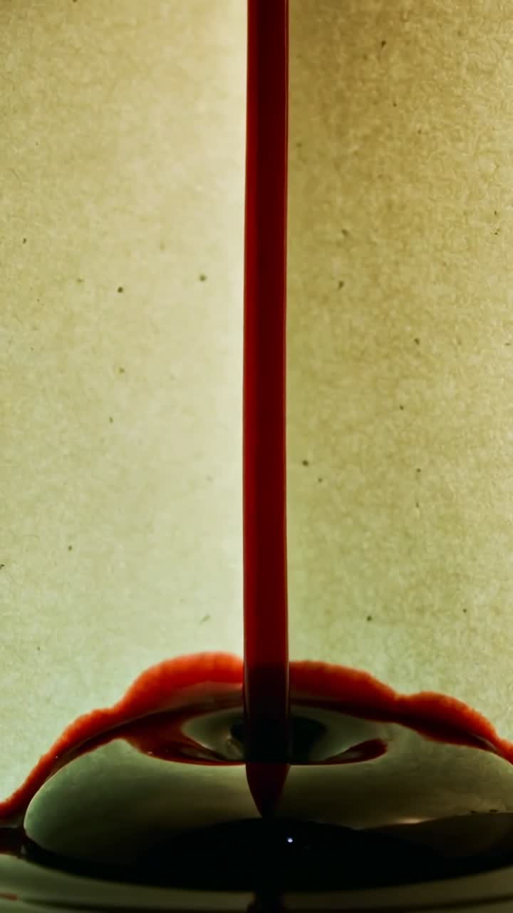 Video verticale: Ugello che gocciola alimentando una colonna di liquido rosso, creando una corona di goccioline su uno sfondo beige.