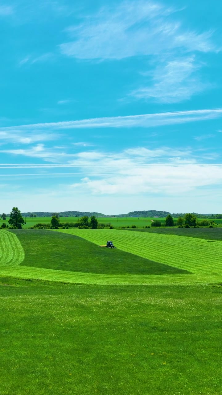 Tomada vertical cinematográfica de máquinas agrícolas trabalhando em um campo verde vibrante sob um céu azul claro. Destacando práticas agrícolas sustentáveis e a beleza da paisagem rural.