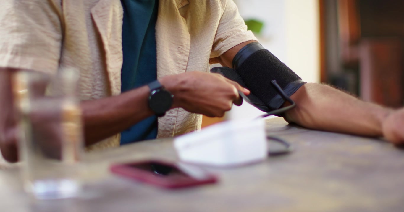 Homem afro-americano colocando manguito no braço na mesa, pressionando monitor branco, verificando a pressão arterial.