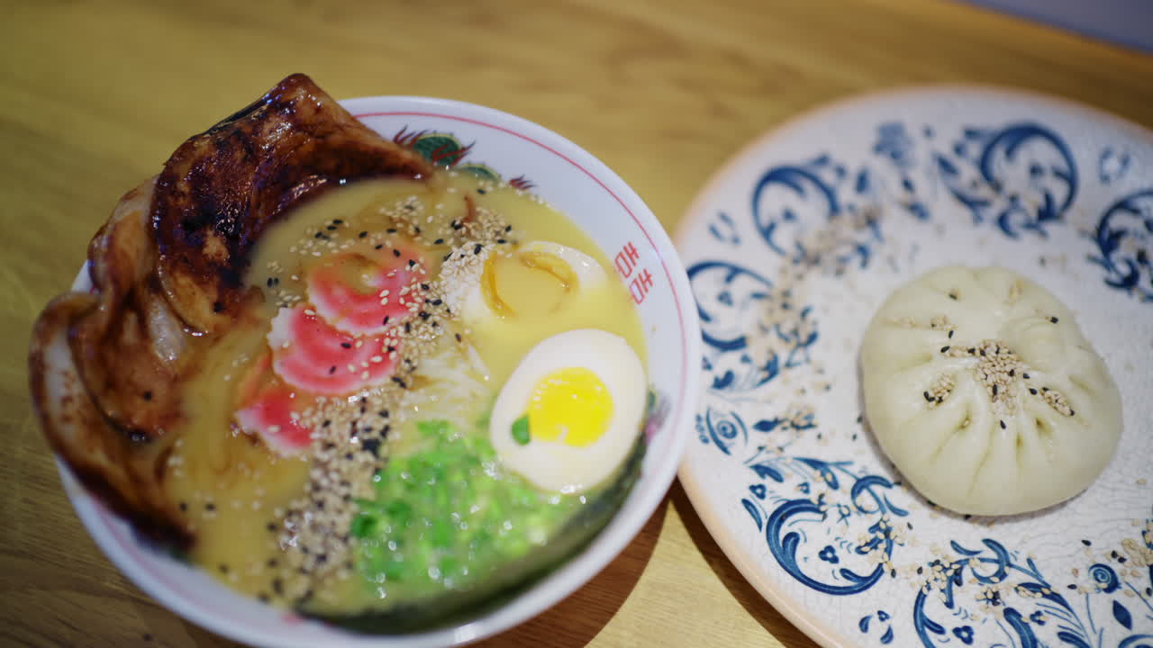 チャーシュー、半熟卵、麺、青菜、ごまが乗ったラーメン鉢と、隣に置かれた蒸しパン。