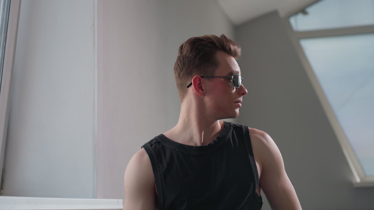 Stylish teen with geometric neck tattoo sits in modern loft by large window, smiles as he lifts sleek dark sunglasses to eyes, soft daylight illuminating his confident pose and casual black outfit