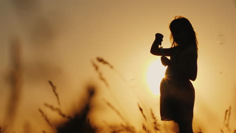 Silhouette-Eines-Mädchens,-Das-Bei-Sonnenuntergang-Mit-Seifenblasen-Im-Hohen-Gras-Spielt