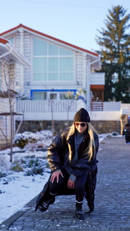 woman poses in front of the camera. model wearing warm winter clothes and sunglasses. cottages at backdrop. vertical video.