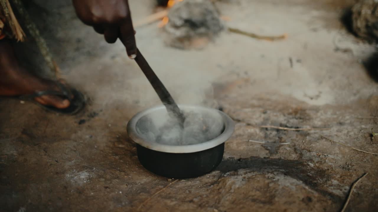 African lady stirring smoking coffee beans in metal pot on ground - traditional method of roasting in Uganda