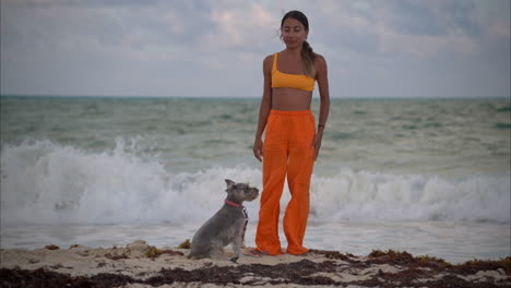 thin sexy hispanic latin girl in orange clothes petting her grey schnauzer dog at the beach with a wave braking in the back