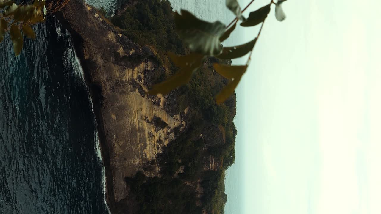 cámara lenta vertical que revela una toma de la isla de nusa penida desde bali en indonesia con vista a otra isla con acantilados y árboles durante las olas tranquilas en un día de mañana ventoso