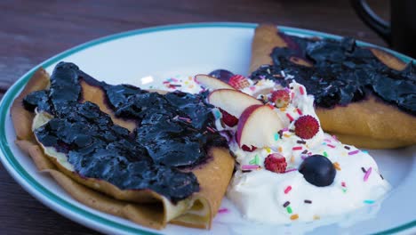 crepas caseras rellenas de queso crema, cubiertas con mermelada de arándanos, servidas con crema batida y frutas frescas como manzanas, fresas y arándanos