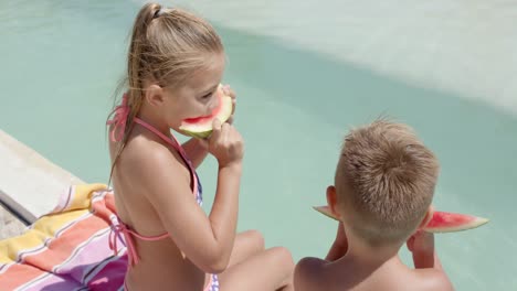 happy caucasian siblings eating watermelon at swimming pool at beach house