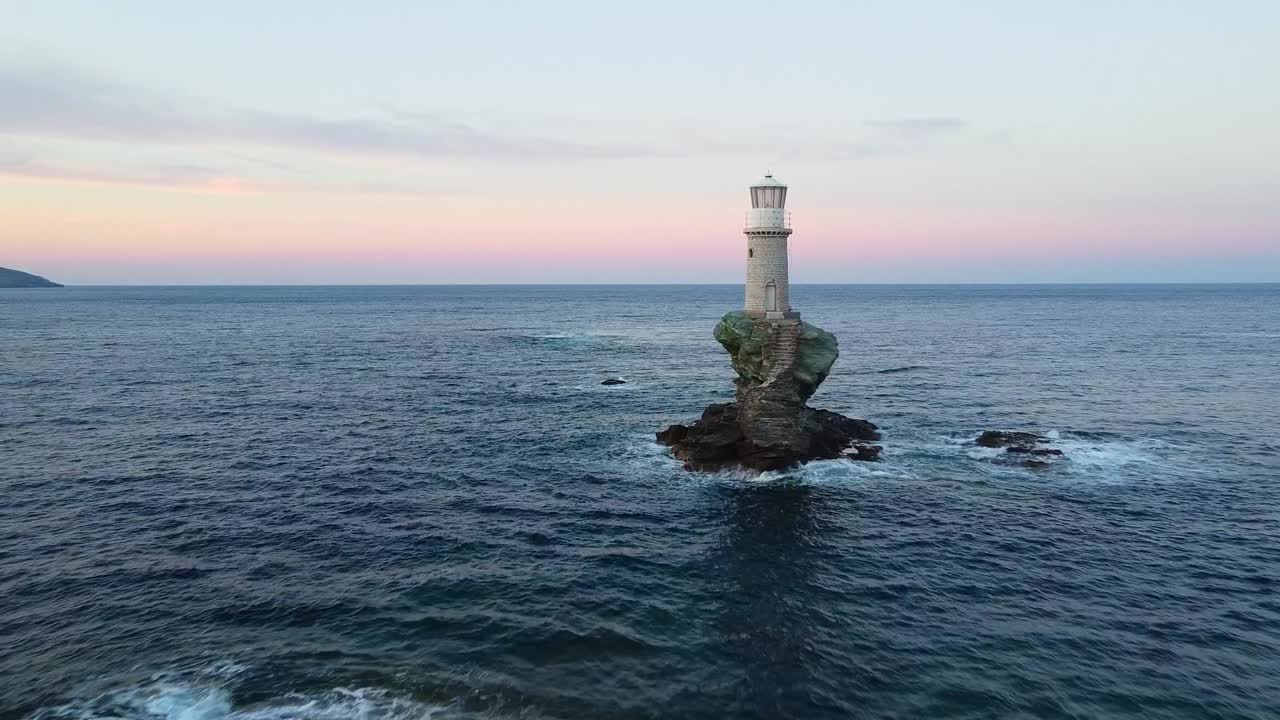 Tourlitis Lighthouse with spiral staircase on a rocky islet from the port of Chora at sunset, on Greek island of Andros, Aegean Sea, Drone shot