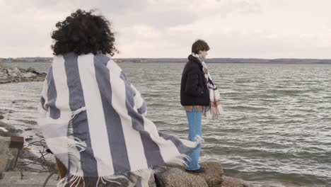 Two-friends-in-winter-clothes-throwing-pebbles-to-the-water-on-a-seashore-on-a-cloudy-day