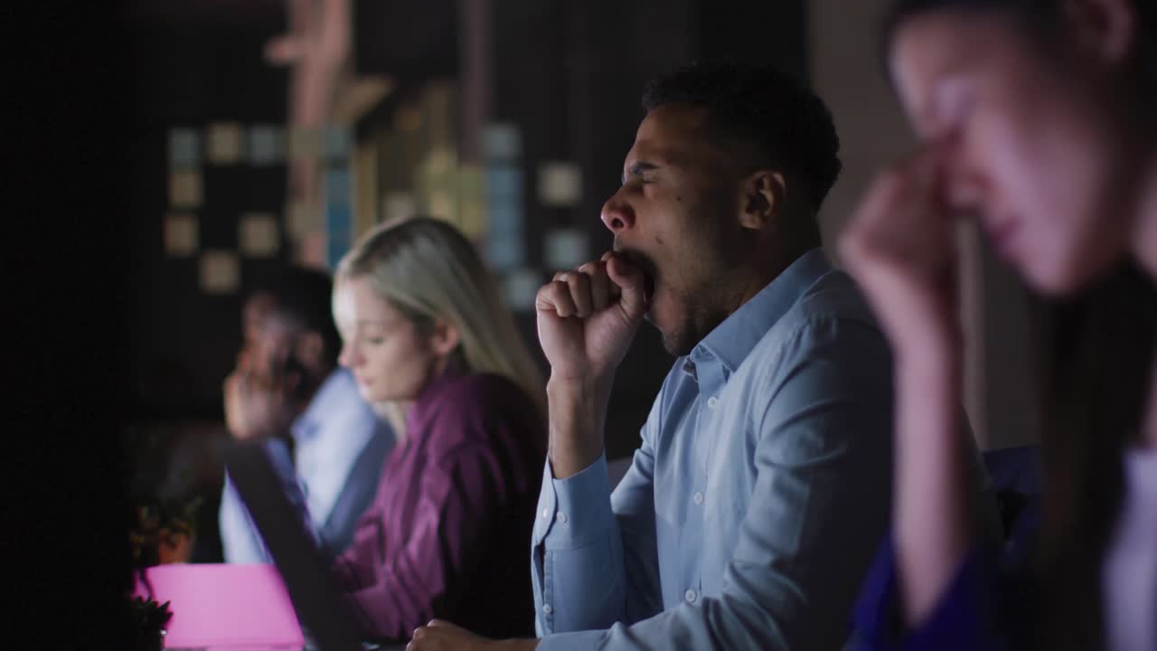Video of tired biracial businessman sitting at desk working at night with colleagues in office