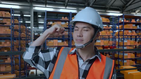 primer plano de un ingeniero masculino asiático con casco de seguridad de pie en el almacén con estantes llenos de productos de entrega. flexionando su bíceps y sonriendo a la cámara en el almacenamiento