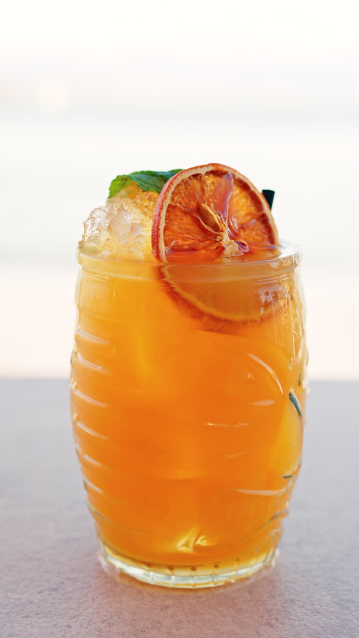 Close up of an orange cocktail on a table with a blurred view of the sea on the background. Vertical