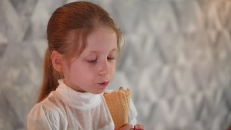 close up of young girl with long brown hair wearing white blouse enjoying ice cream cone indoors, savoring sweet treat with content look, standing against soft patterned wall background