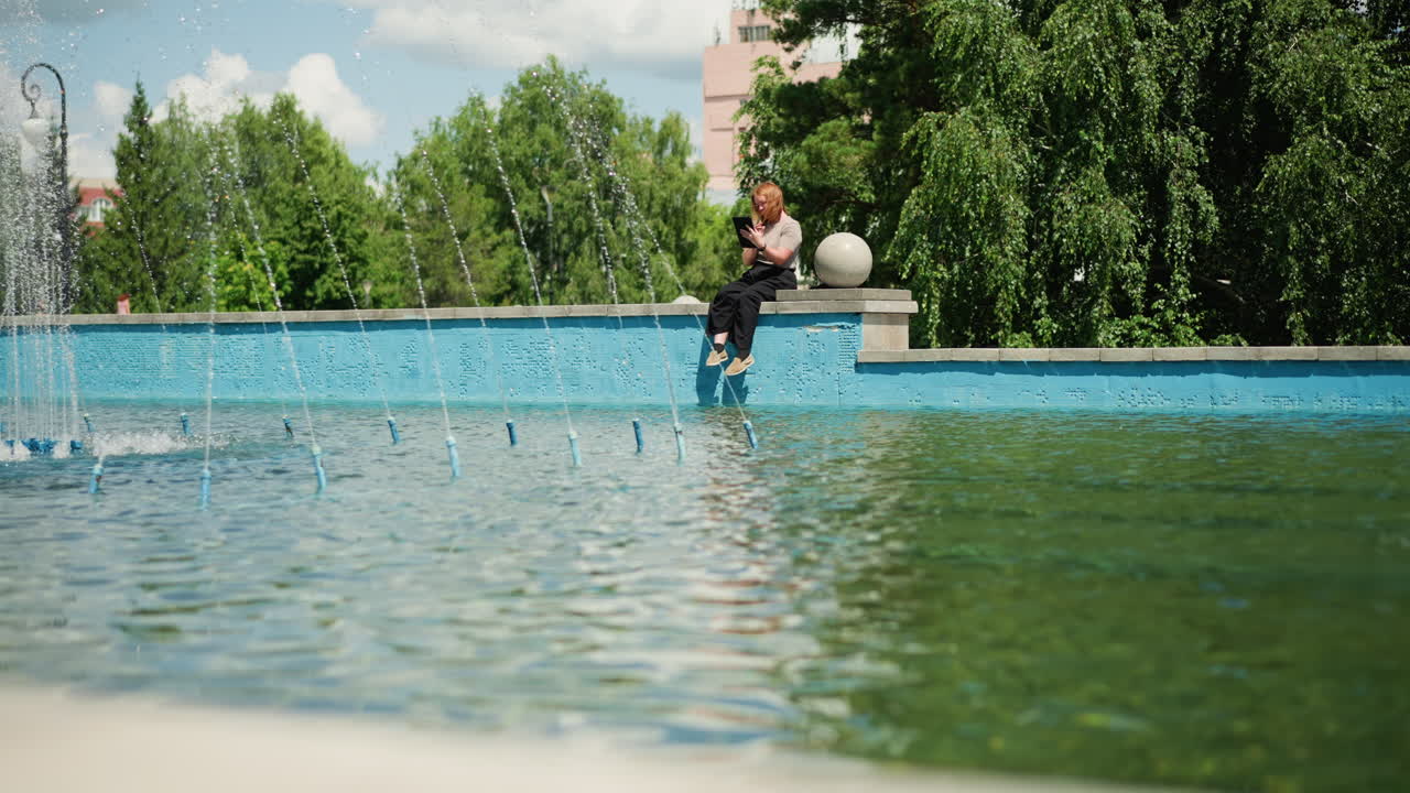 Tech lady sitting by fountain using tablet under bright sunlight, surrounded by green trees and calm water, enjoying peaceful outdoor work moment with focus