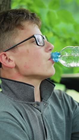 dos hermanos jóvenes descansando bajo un árbol con uno de ellos bebiendo agua mientras el otro inclina la cabeza, están tomando un descanso de un paseo en bicicleta, con bicicletas estacionadas cerca rodeadas de vegetación