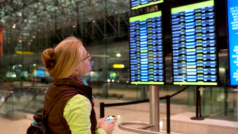 foto lateral de una mujer rubia apuntando a la pantalla de información de vuelo mientras sostiene su teléfono inteligente en un aeropuerto