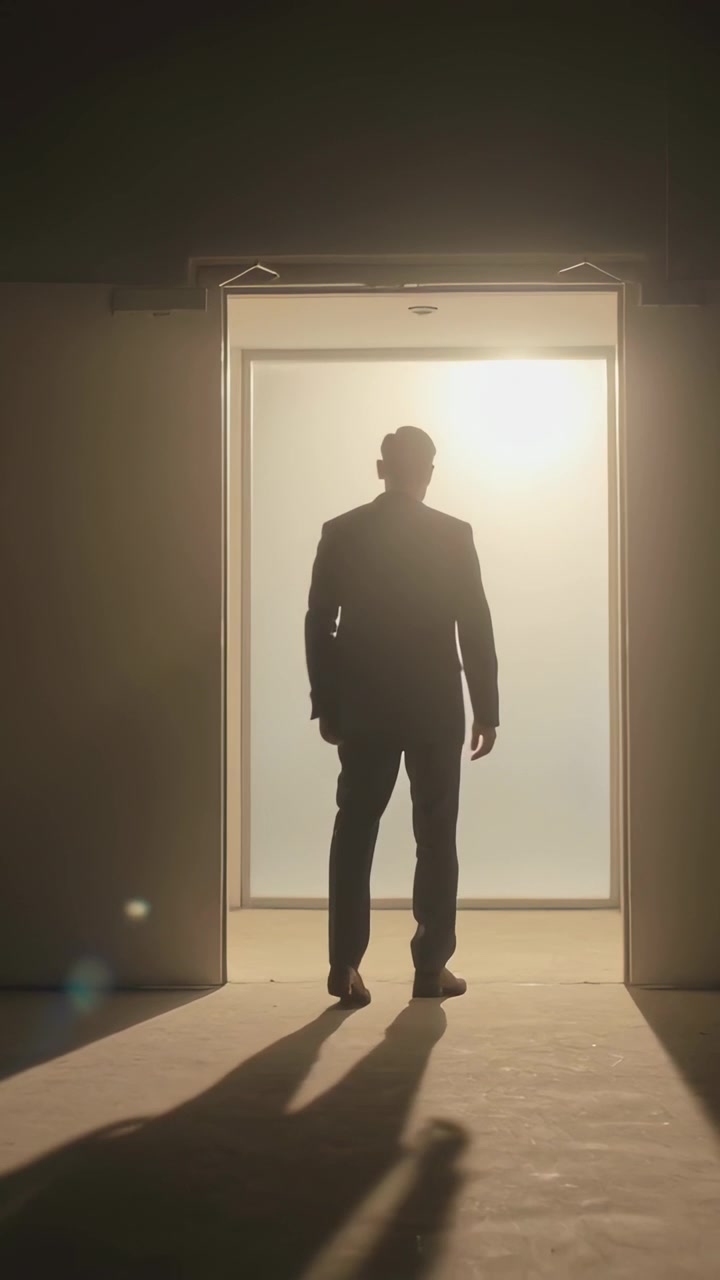 Video verticale: Uomo in abito scuro che cammina ed entra in una porta retroilluminata in una stanza in penombra, la luce lo attira, fermandosi.
