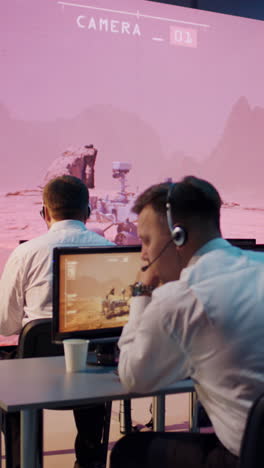 young female employee in headset typing on computer keyboard and watching video from mars research vehicle while sitting at desk near colleagues in control room. vertical shot