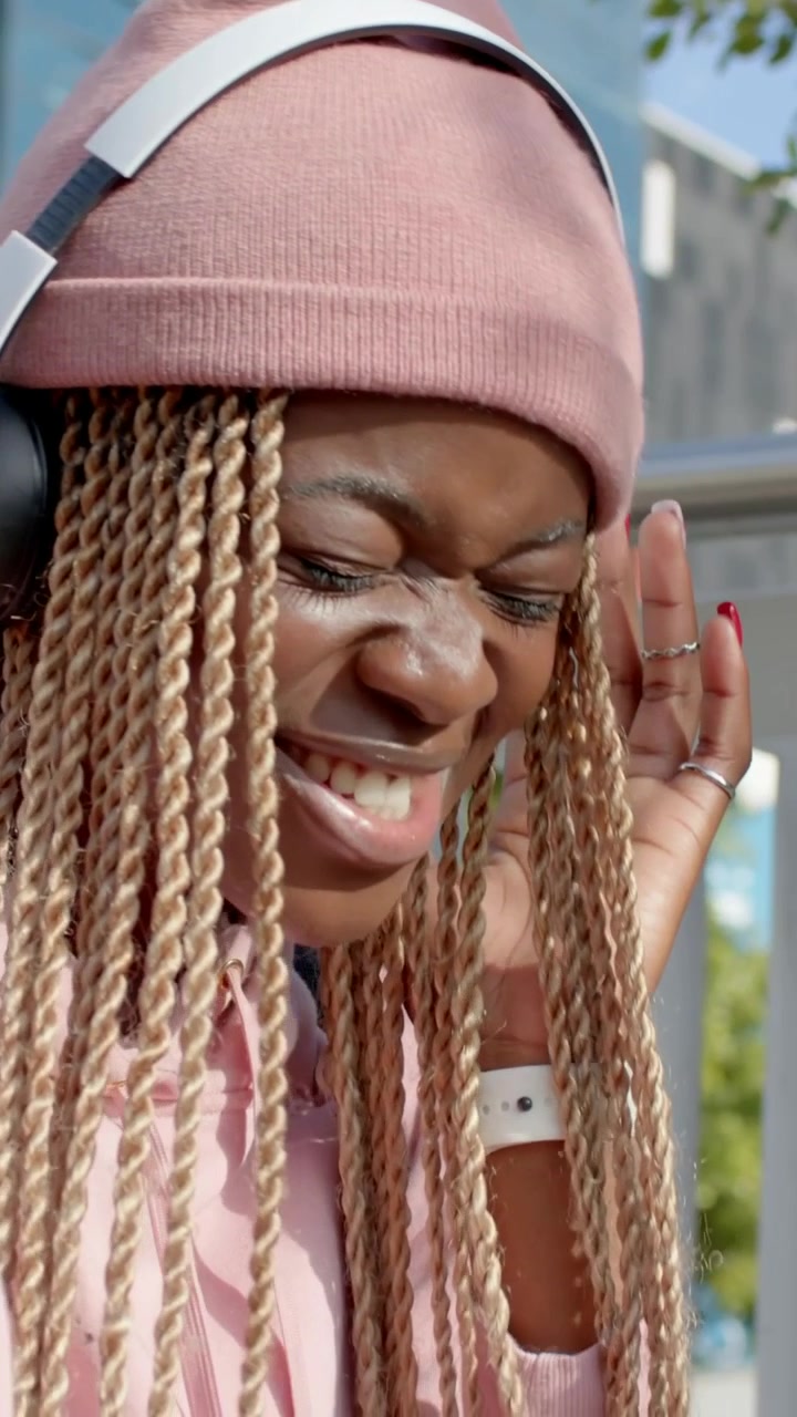 Vertical video: Pressing earcups, woman in pink beanie singing by glass building as song changes