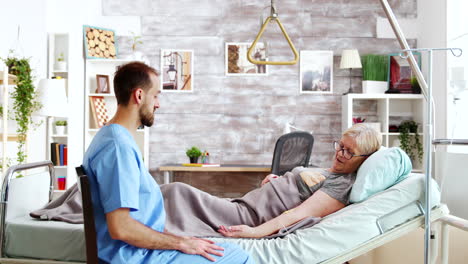 male assistant taking a seat near hospital bed of ill lady lying in nursing home