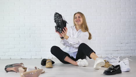 Cheerful young woman sitting on the floor and showing various footwear. Shoe presentation at the white wall backdrop.