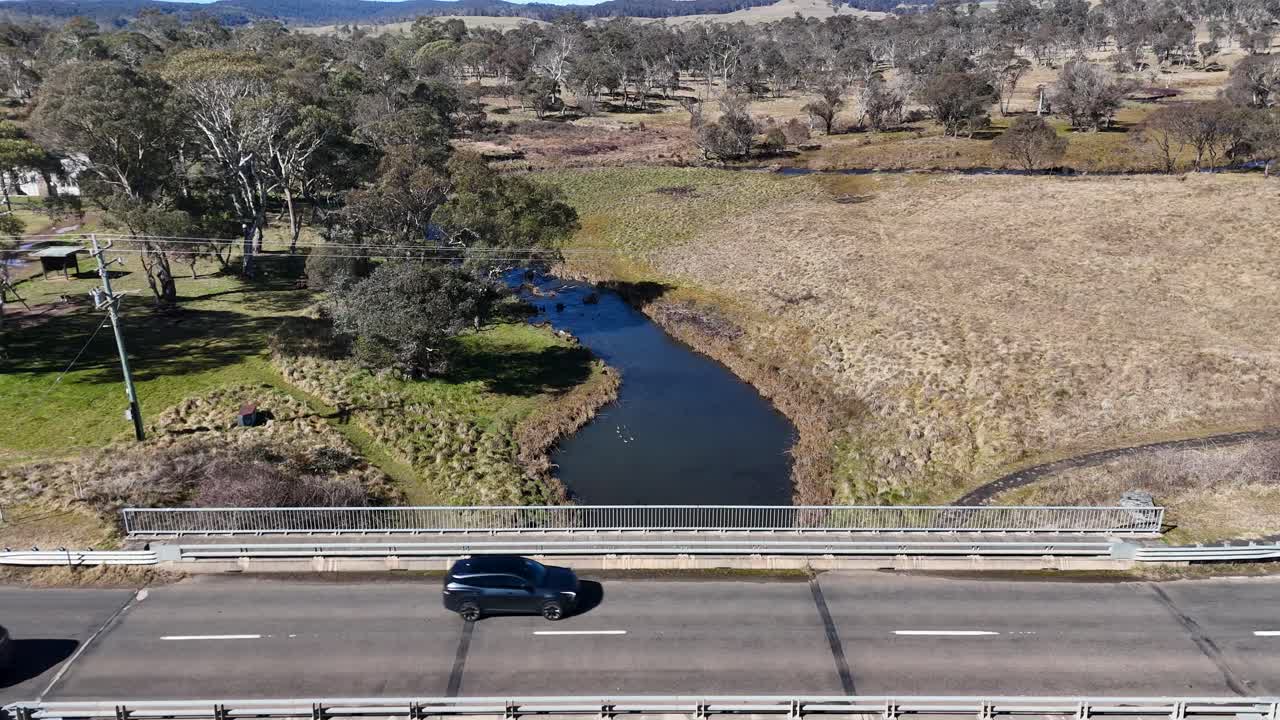 Drone footage tracks vehicles driving across a concrete bridge spanning a winding stream in a grassy, rural landscape under bright daylight