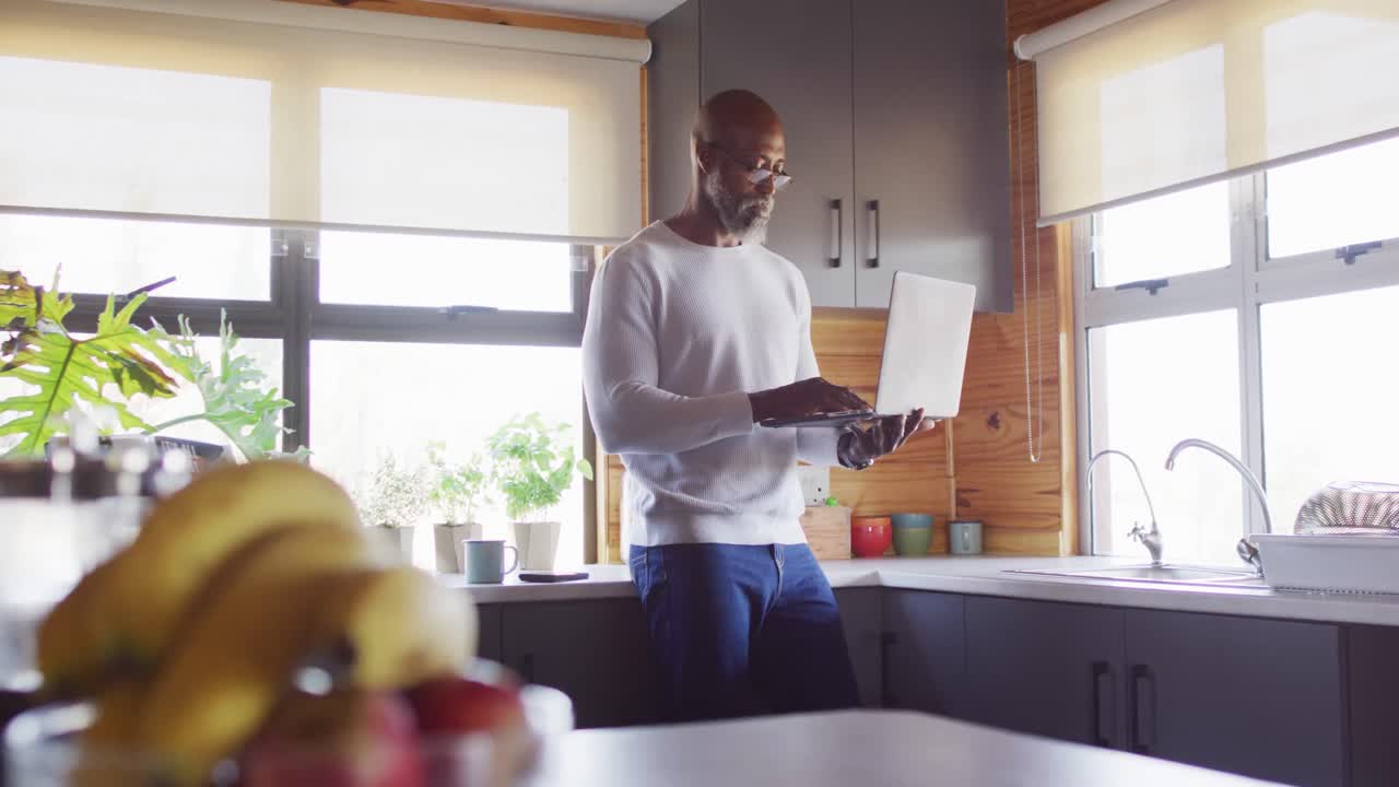 Happy senior african american man in log cabin, standing in kitchen and using laptop, slow motion