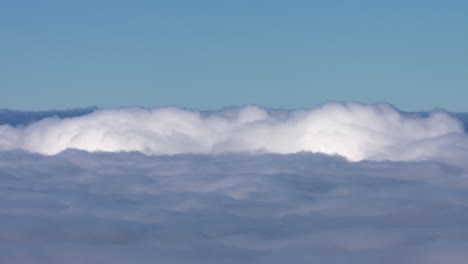 landscape shot above low lying white fluffy clouds, showing the clouds rolling, bright blue sky above