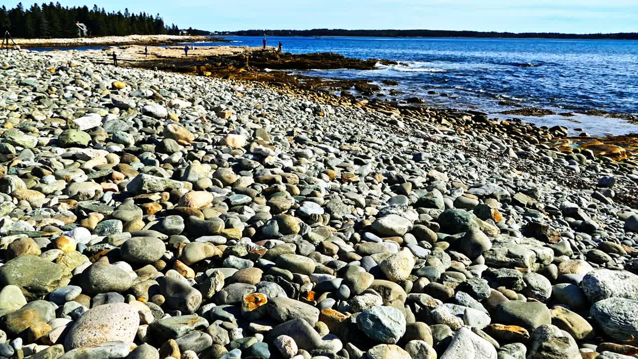 Naturskøn udsigt over en robust, stenet strand, der møder det dybblå hav. Blide bølger skvulper mod kysten under en klar himmel på en solrig dag.