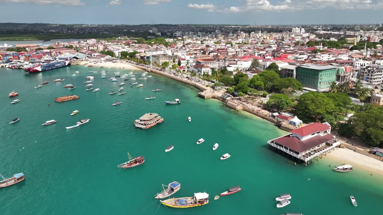 Ripresa cinematografica in avvicinamento con drone che sorvola le imbarcazioni in direzione dei giardini di Forodhani a Stone Town, Zanzibar, in una soleggiata giornata estiva. L'acqua turchese, la vita costiera e lo skyline storico creano un'atmosfera vivace.