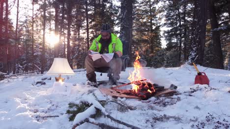 man sitting on a chair next to a fire and lamp in a forest covered with snow