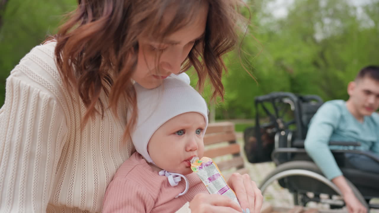 Caring Family Sharing Snack Outdoors, Warm Community Scene With Family Feeding In Park, Caucasian Family Enjoying Outdoor Meal In Accessible Park Environment With Wheelchair Present