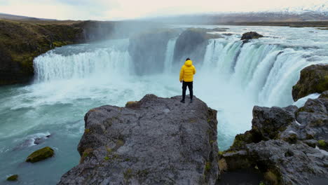 vista de un hombre de pie en un acantilado junto a la cascada godafoss en islandia - retroceso aéreo