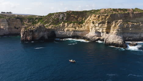 Aerial-view-around-a-boat-on-the-steep,-rocky-coastline-of-the-sunny-Malta-island