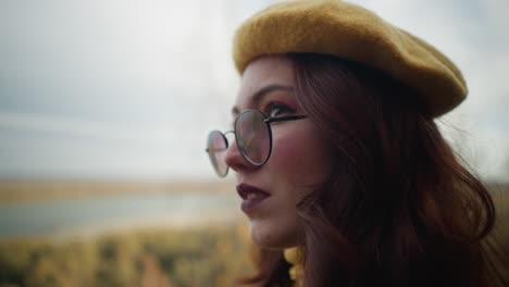 close-up of stylish woman in yellow beret and glasses, gazing into distance with soft smile, autumn trees and river create blurred background, adding warmth and depth to thoughtful expression