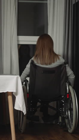 woman moves closer to window spinning big wheels of wheelchair. housewife looks out dark window while spending weekend evening in kitchen backside view