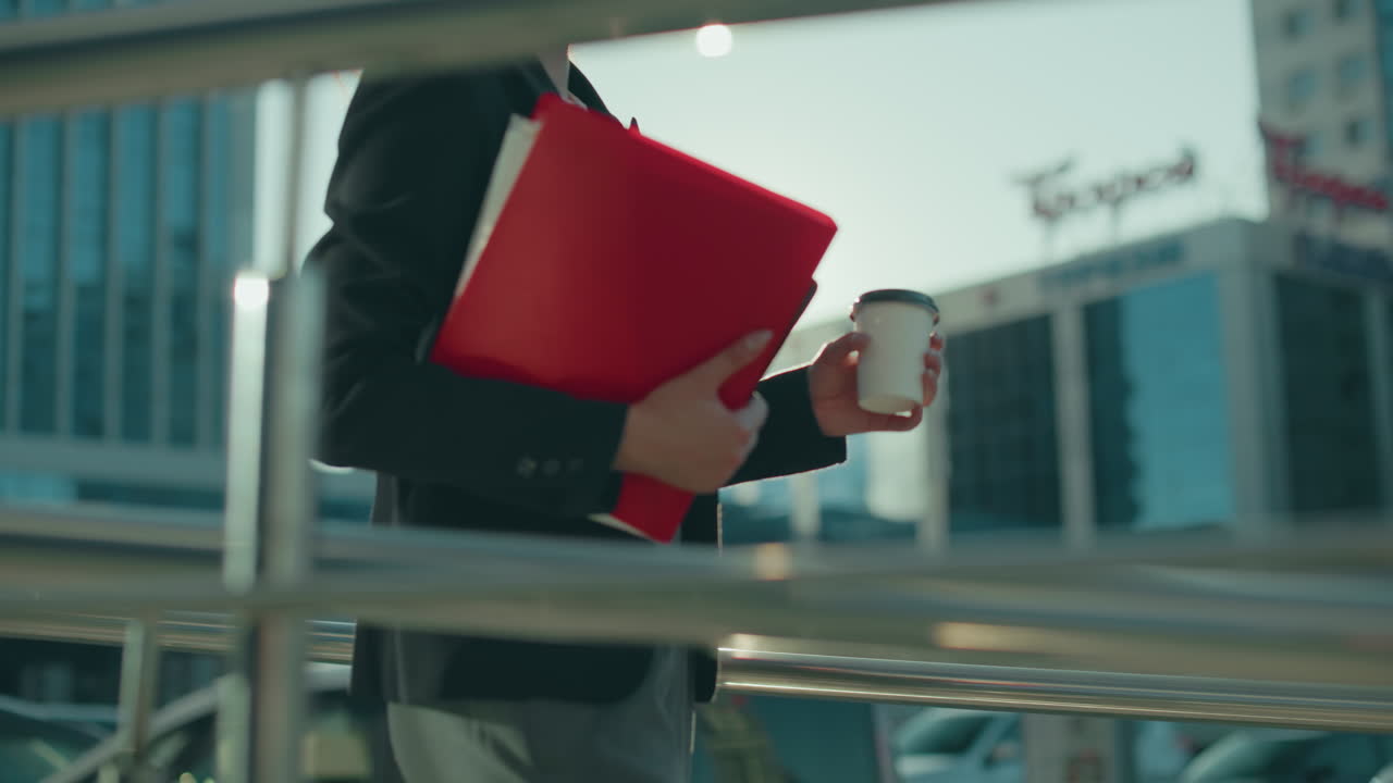 Close up of iron railing framing view of woman walking outdoors holding red folder and takeaway coffee cup, modern glass office buildings and parked cars visible in background