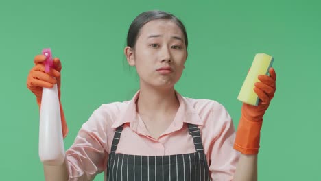 close up of tired asian female housekeeper with an apron looking at spray and kitchen sponge in hands then shaking her head in the green screen studio