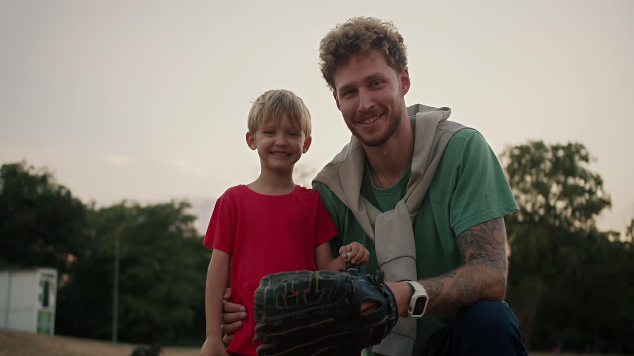 Portrait of a Happy little blond boy with blue eyes in a red T-shirt who stands with his dad with curly hair with stubble in a Green T-shirt and wearing a baseball glove in the park