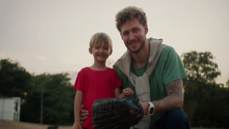 Portrait-of-a-Happy-little-blond-boy-with-blue-eyes-in-a-red-T-shirt-who-stands-with-his-dad-with-curly-hair-with-stubble-in-a-Green-T-shirt-and-wearing-a-baseball-glove-in-the-park