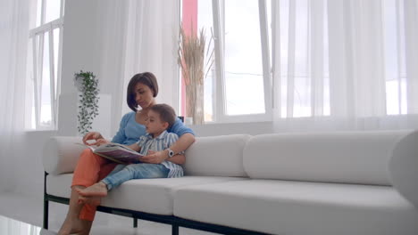 Una-Joven-Madre-Con-Un-Niño-Leyendo-Un-Libro-Sentado-En-Un-Interior-Blanco-Brillante-De-La-Casa-En-La-Sala-De-Estar-En-El-Sofá