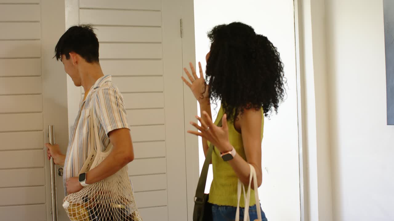 Diverse couple arriving home with groceries, man holding mesh bag, woman gesturing, closing door