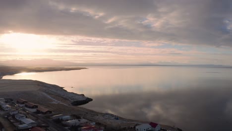 sensational sunset aerial view of icelandic harbor town by sea on calm winter day, snowy mountains in background, drone flying forward above ocean, cloudy day