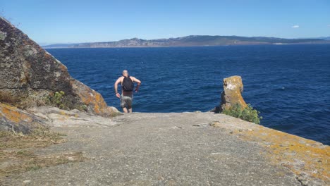 Hombre-Sin-Ropa-Con-Mochila-Viene-Corriendo-Para-Ayudar-Al-Acantilado-Del-Mar-Con-La-Costa-Al-Fondo-Un-Día-Soleado-Sin-Nubes,-Tiro-Bloqueado-Bajo,-Islas-Cíes,-Pontevedra,-España