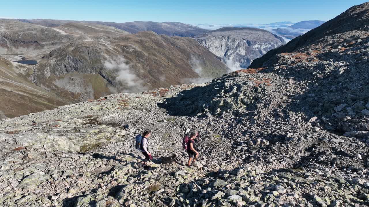 Two fit brunette women walk on a rocky ground toward Bakkanosi, followed by a low-altitude sideways drone in summer