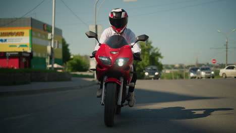 un motociclista en un casco monta una motocicleta roja con un faro encendido a través de una calle de la ciudad, los coches están esperando para girar, y los camiones están estacionados en el lado opuesto