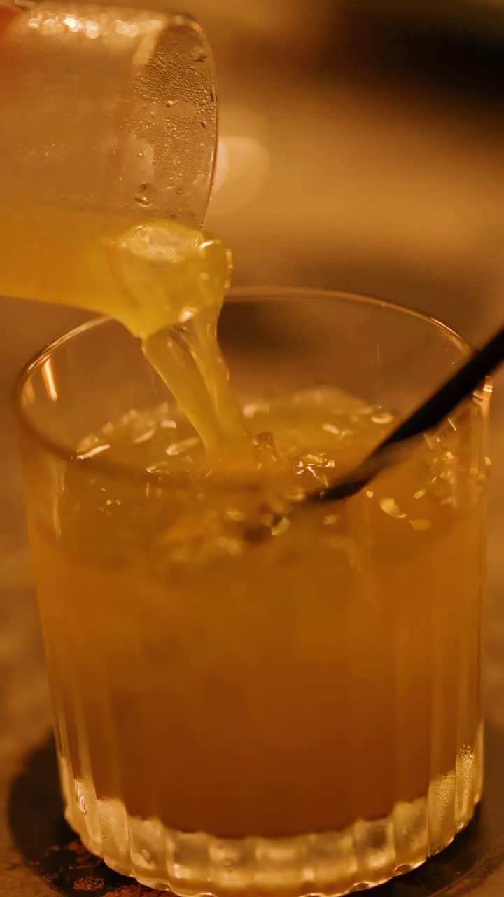 Un cocktail viene versato in un bicchiere con ghiaccio, sotto una luce calda e soffusa, creando un'atmosfera accogliente.