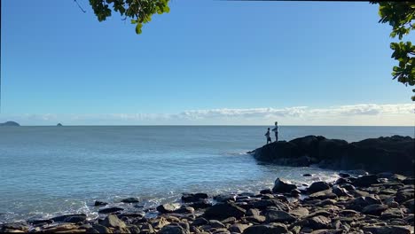 a pair of fishermen fishing from the rocks in this north queensland rocky shoreline setting, with gentle lapping waves under a blue summer sky and overhanging trees in the foreground